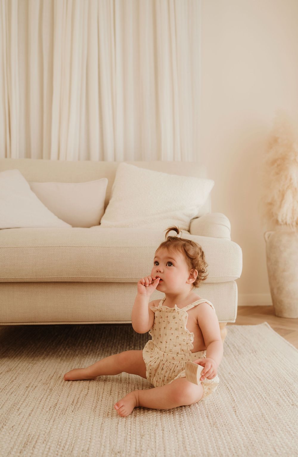 A toddler in a cream lace romper relaxes on a white carpeted floor in a minimalist room setting.