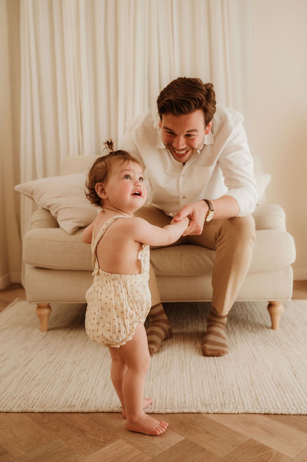 Adult in white shirt sitting on beige couch plays with toddler wearing floral romper on wooden floor in bright room.
