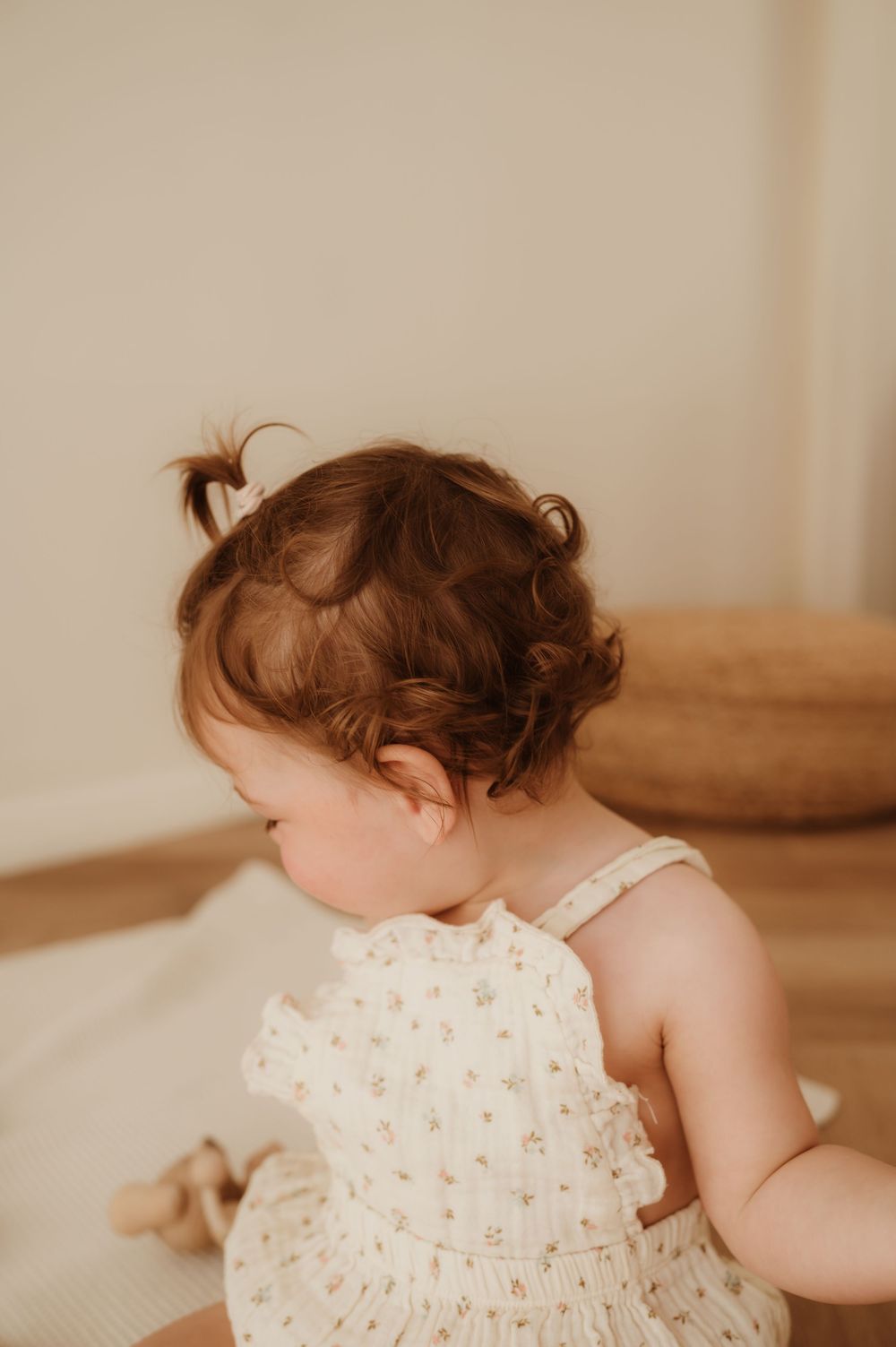 A small toddler wearing a delicate floral dress sits looking down in a warm, sunlit room.