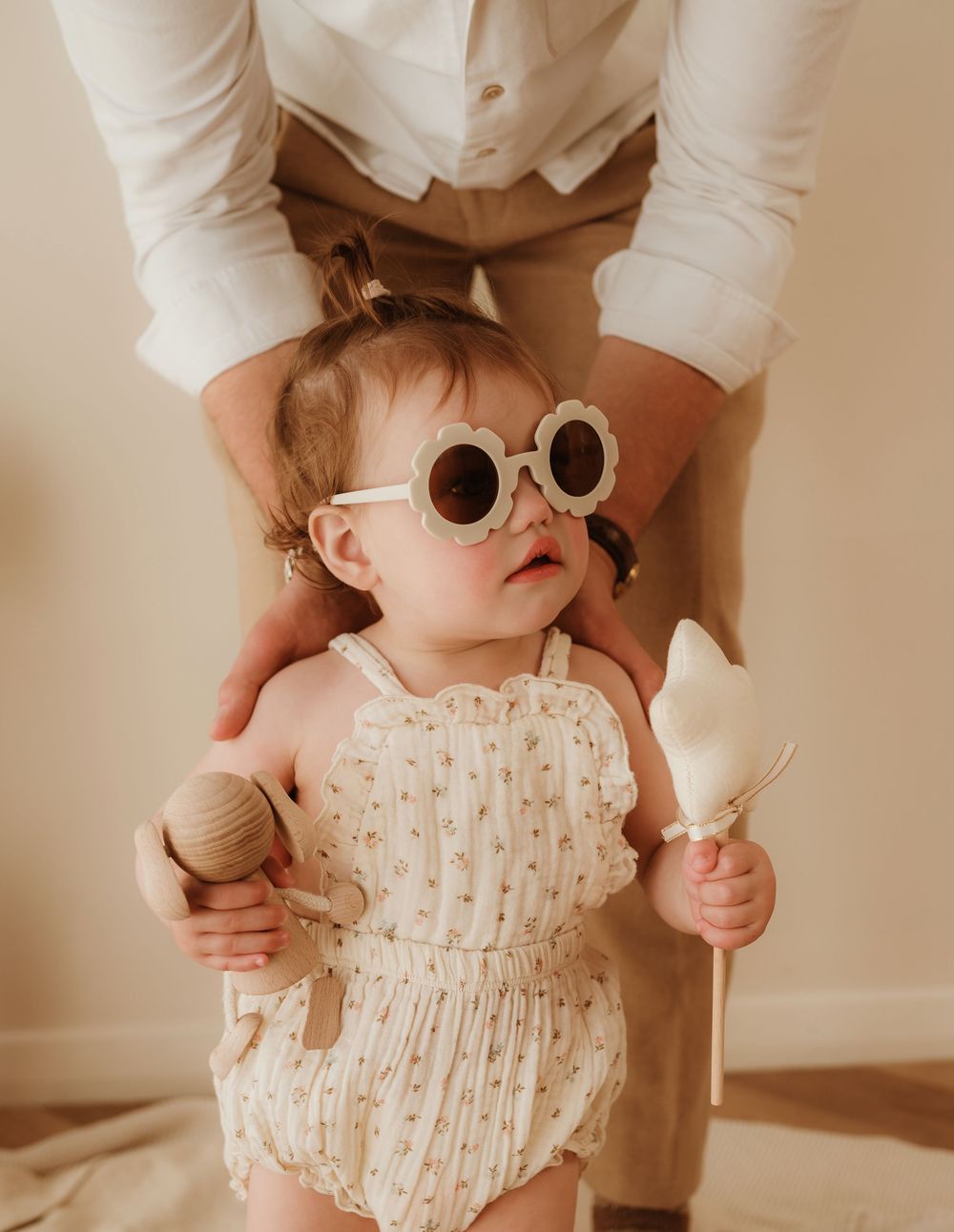 A small child wearing retro sunglasses and a light patterned romper holds a wooden toy and ice cream cone.