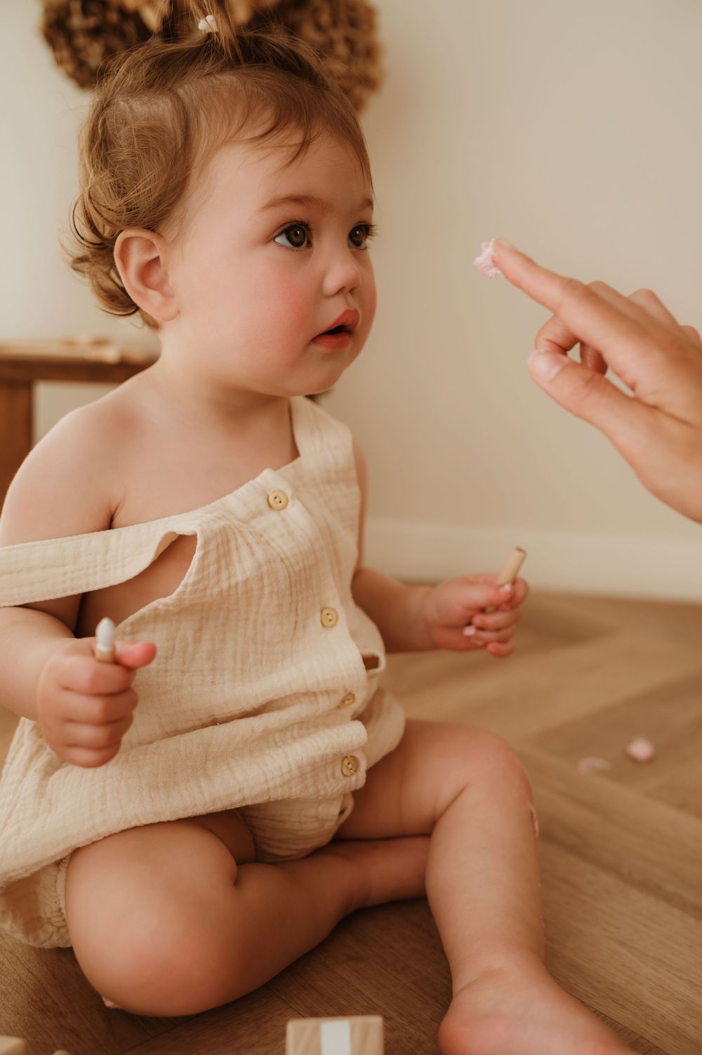Baby in beige cotton romper on wooden floor reaching up while exploring the surroundings.