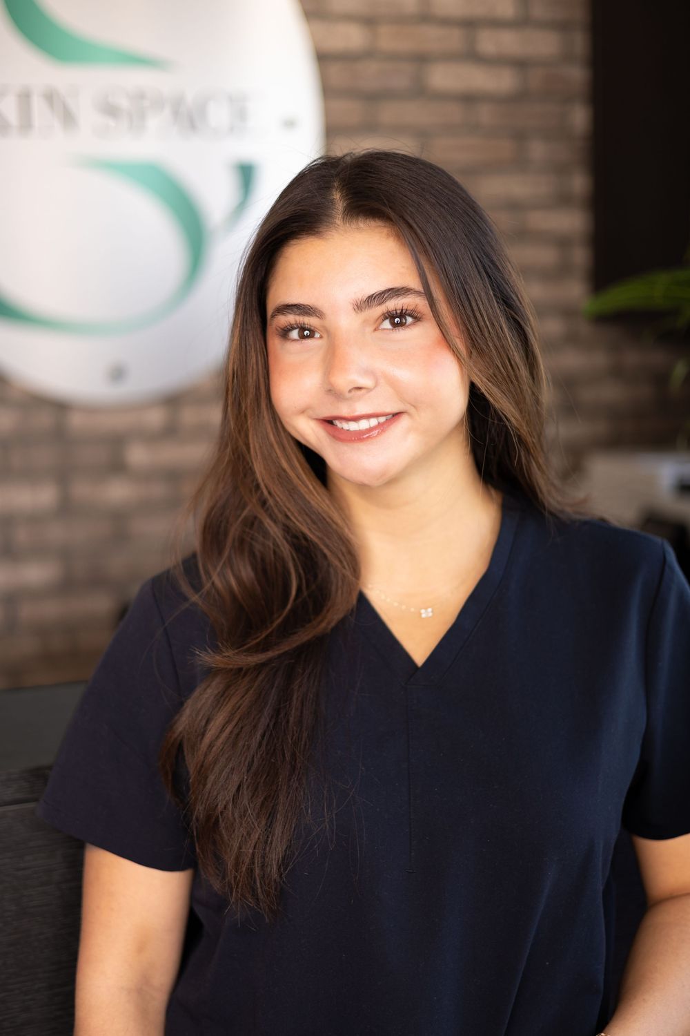 Young woman in navy scrubs smiling at Skin Space clinic, brunette hair, professional skincare staff portrait.