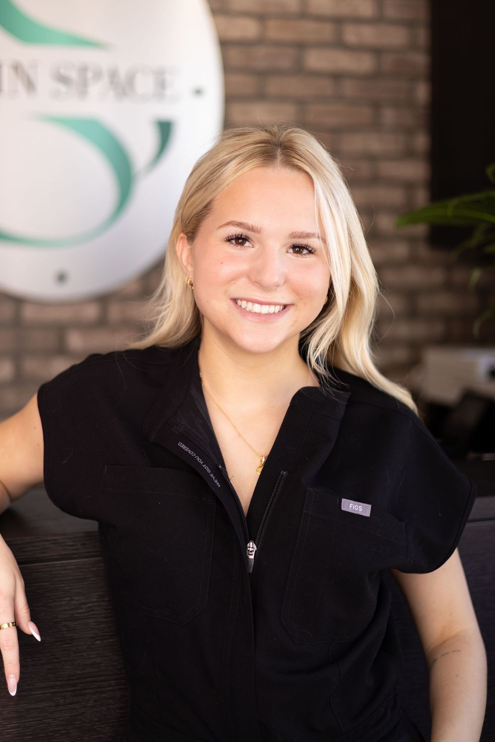 A smiling blonde esthetician in black uniform leans on reception desk at Skin Space spa location.