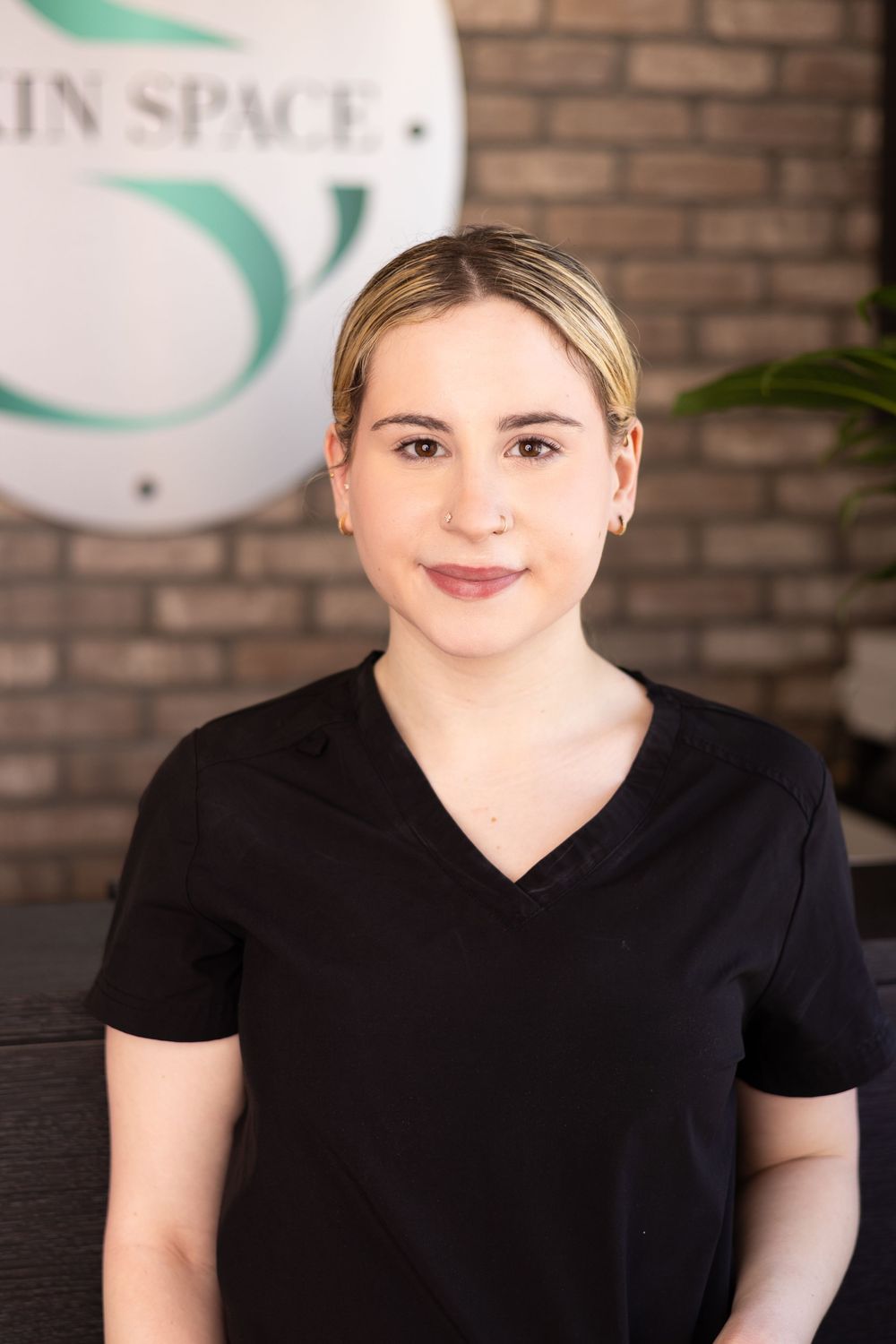 A smiling blonde woman in black scrubs poses professionally in front of a brick wall with a spa logo.