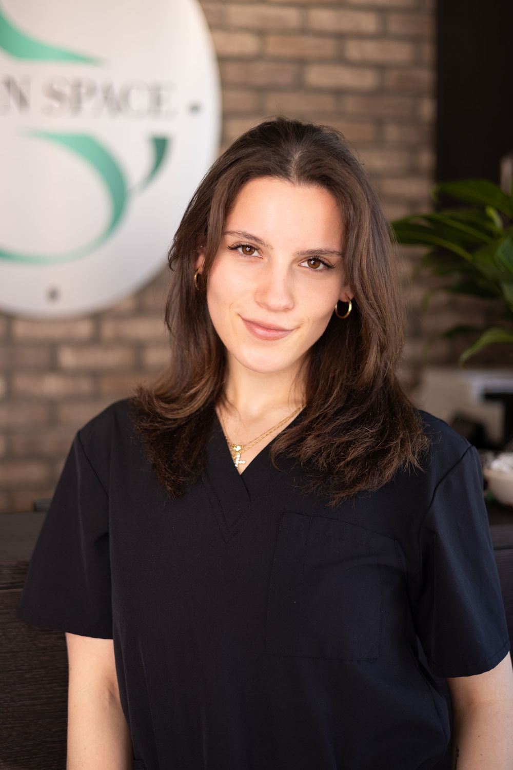 Young woman in black scrubs smiling at a modern spa reception desk with a green logo sign.