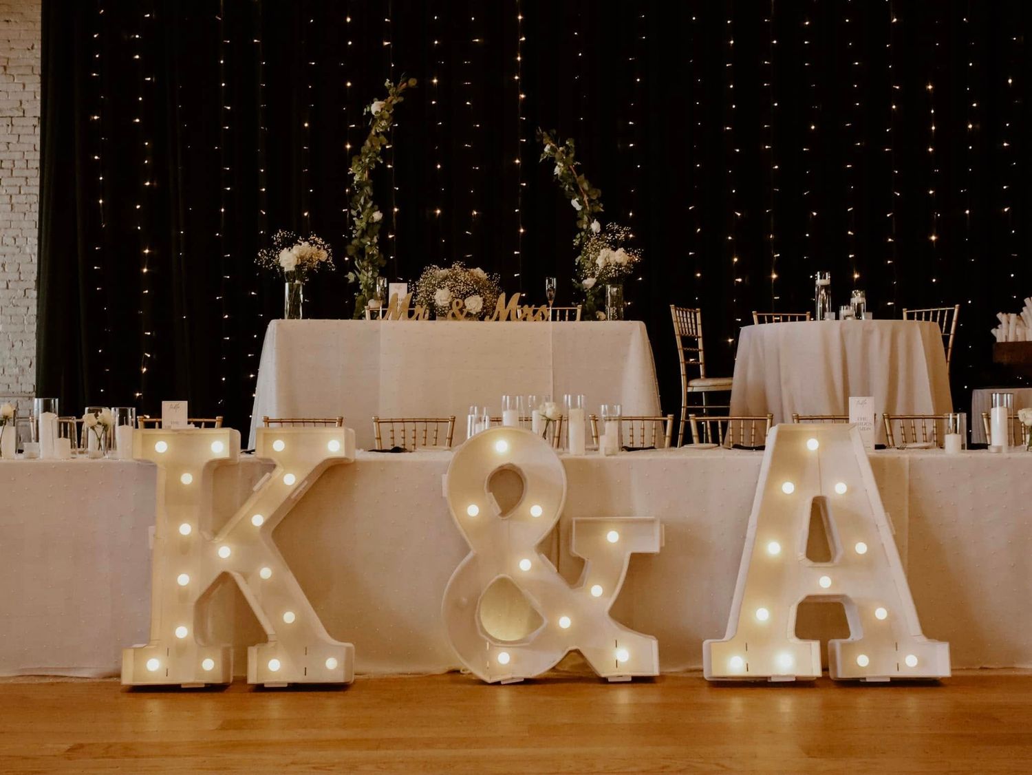 Large white illuminated marquee letters spelling K&A on a table at a wedding reception with twinkle lights and decor.