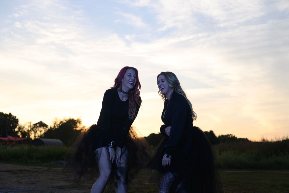 Two people in dark clothing sit outdoors laughing together against a sunset sky backdrop.