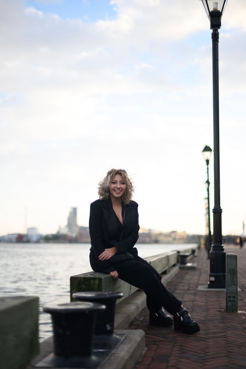 Someone sits on a waterfront bench wearing black clothing against a harbor backdrop with lampposts and city skyline.
