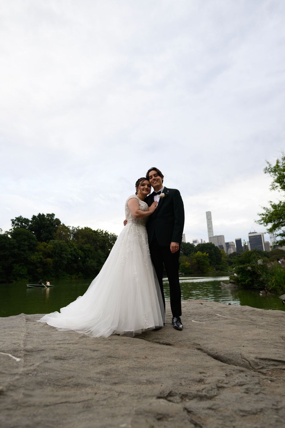 A couple in wedding attire poses on a rock formation overlooking Central Park lake with Manhattan skyline in background.