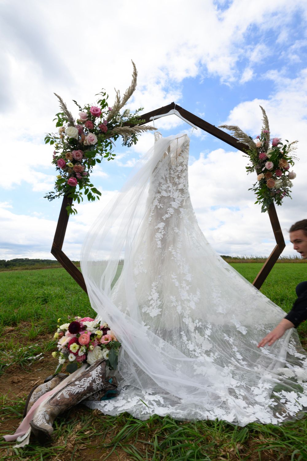A white lace wedding dress displayed on a hexagonal arch decorated with pink and white flowers against a blue sky.