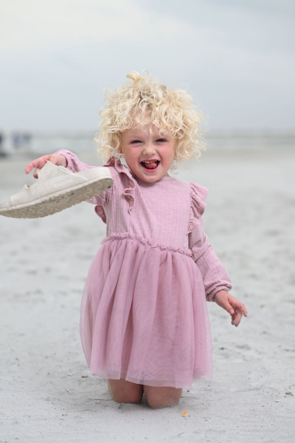 A joyful child wearing a pink dress plays on a sandy beach with curly blonde hair and a bright smile.