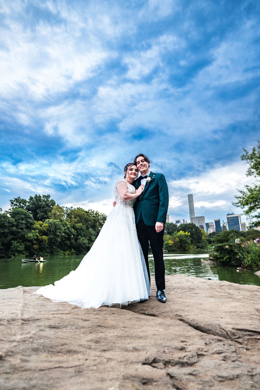 A wedding couple poses on a rocky outcrop in Central Park with the Manhattan skyline and lake in the background.
