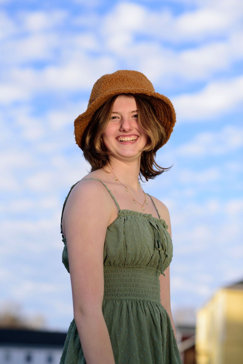 Person in olive green sundress and brown sun hat smiles against blue sky with puffy clouds on sunny day.
