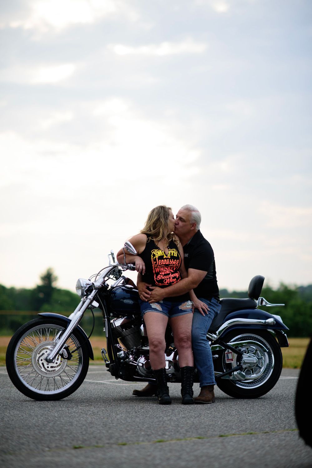 A couple embraces on their parked motorcycle against a cloudy sky backdrop.
