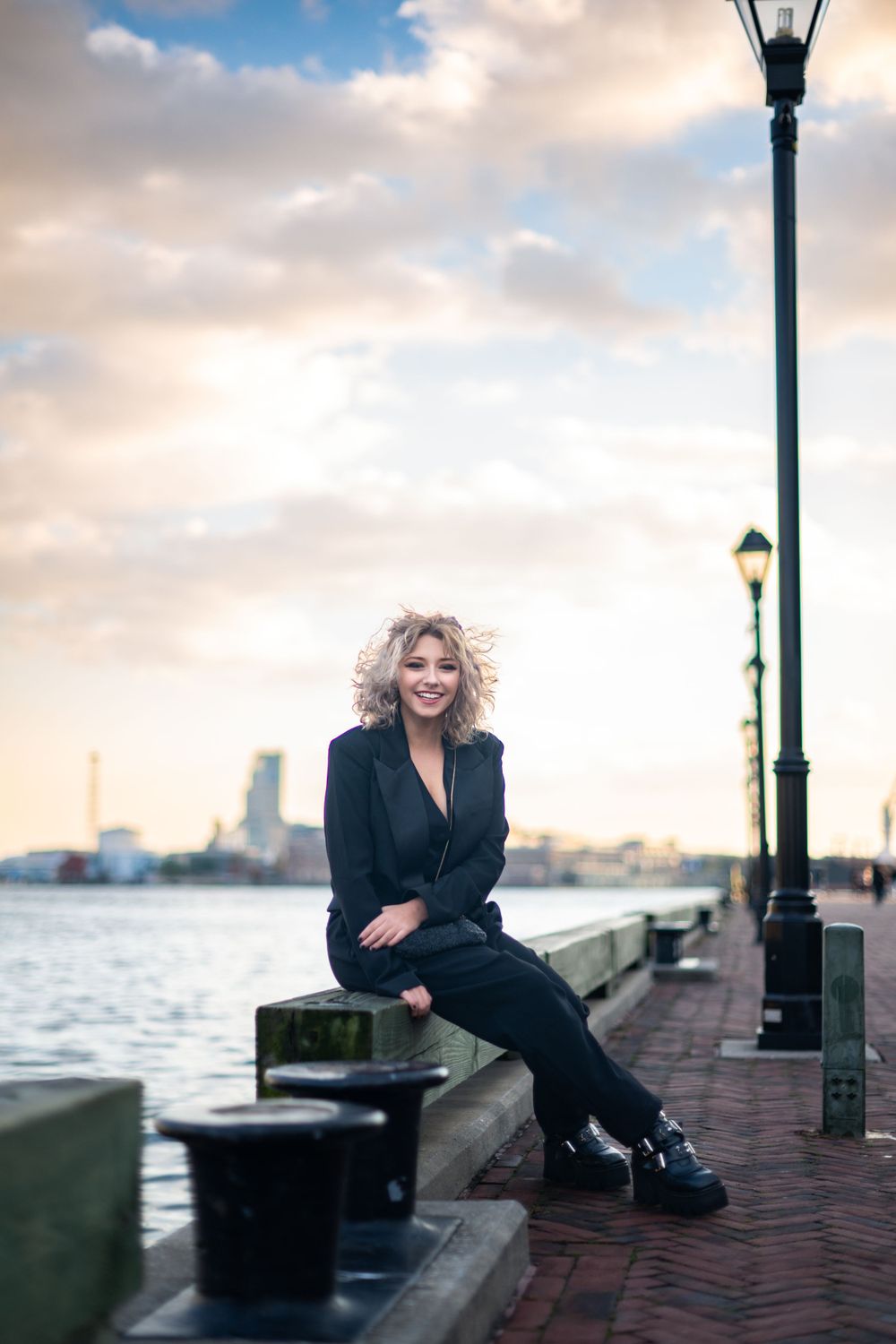 Person in black outfit sits along waterfront boardwalk with city skyline and lampposts in background at dusk.
