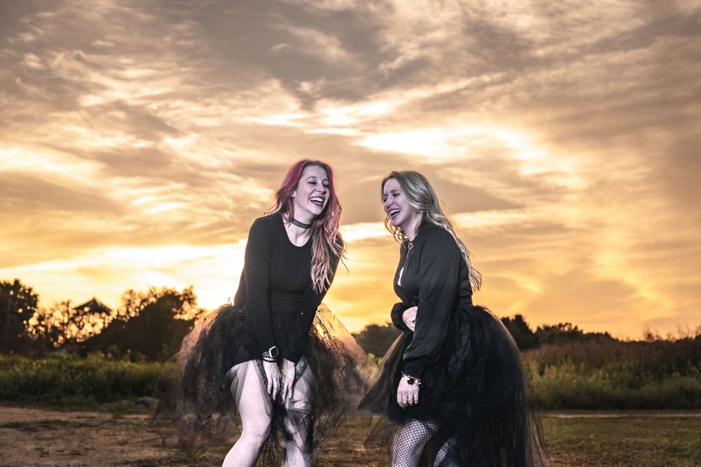Two women in dark flowy dresses dance together against a dramatic golden sunset sky in a rural field.
