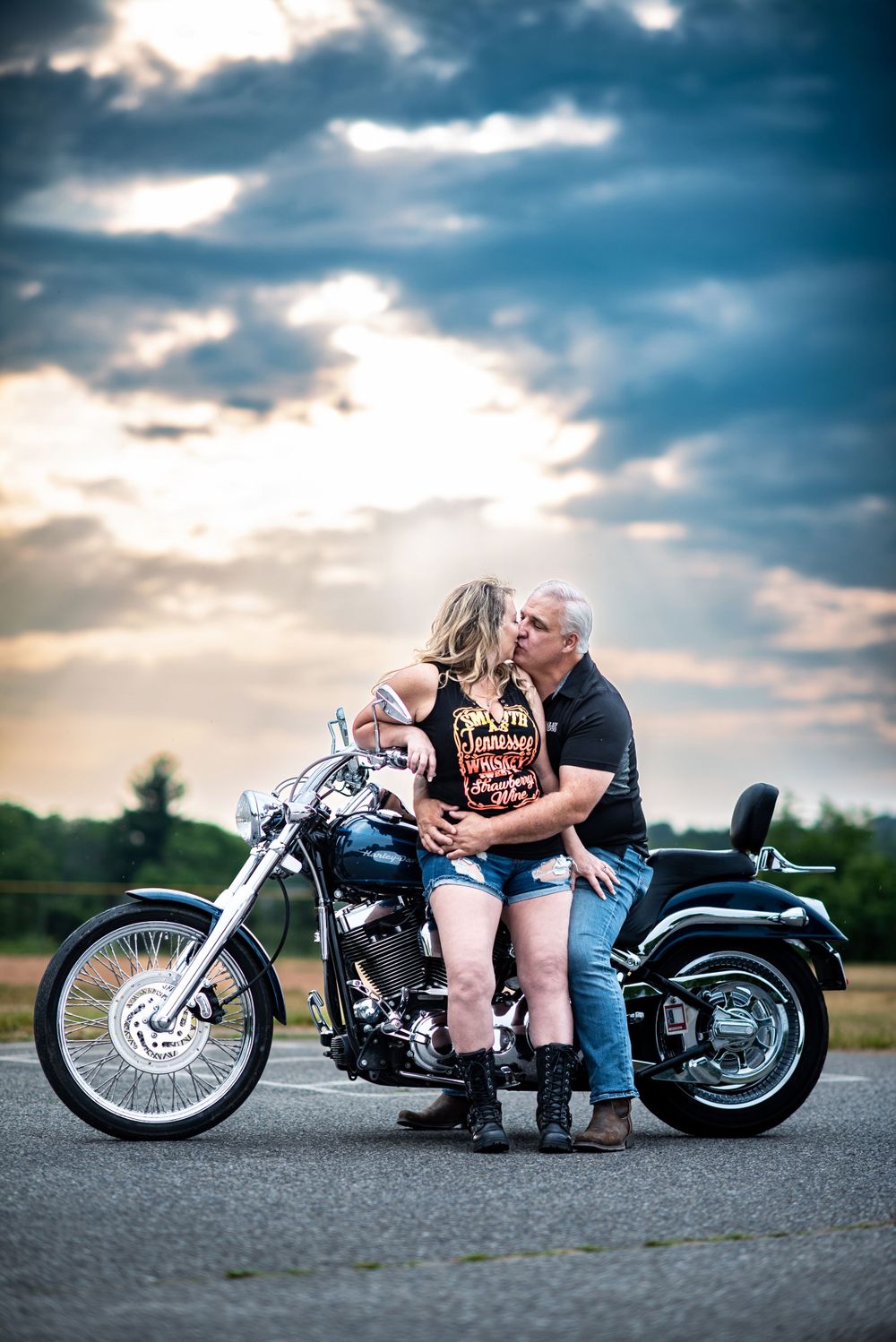 A couple share an intimate moment on a parked motorcycle against a dramatic cloudy sunset sky.