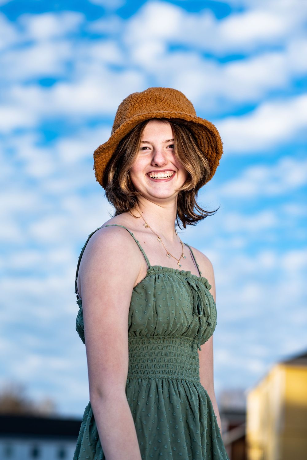 Person in olive green sundress and brown sun hat smiling against blue sky with puffy white clouds.