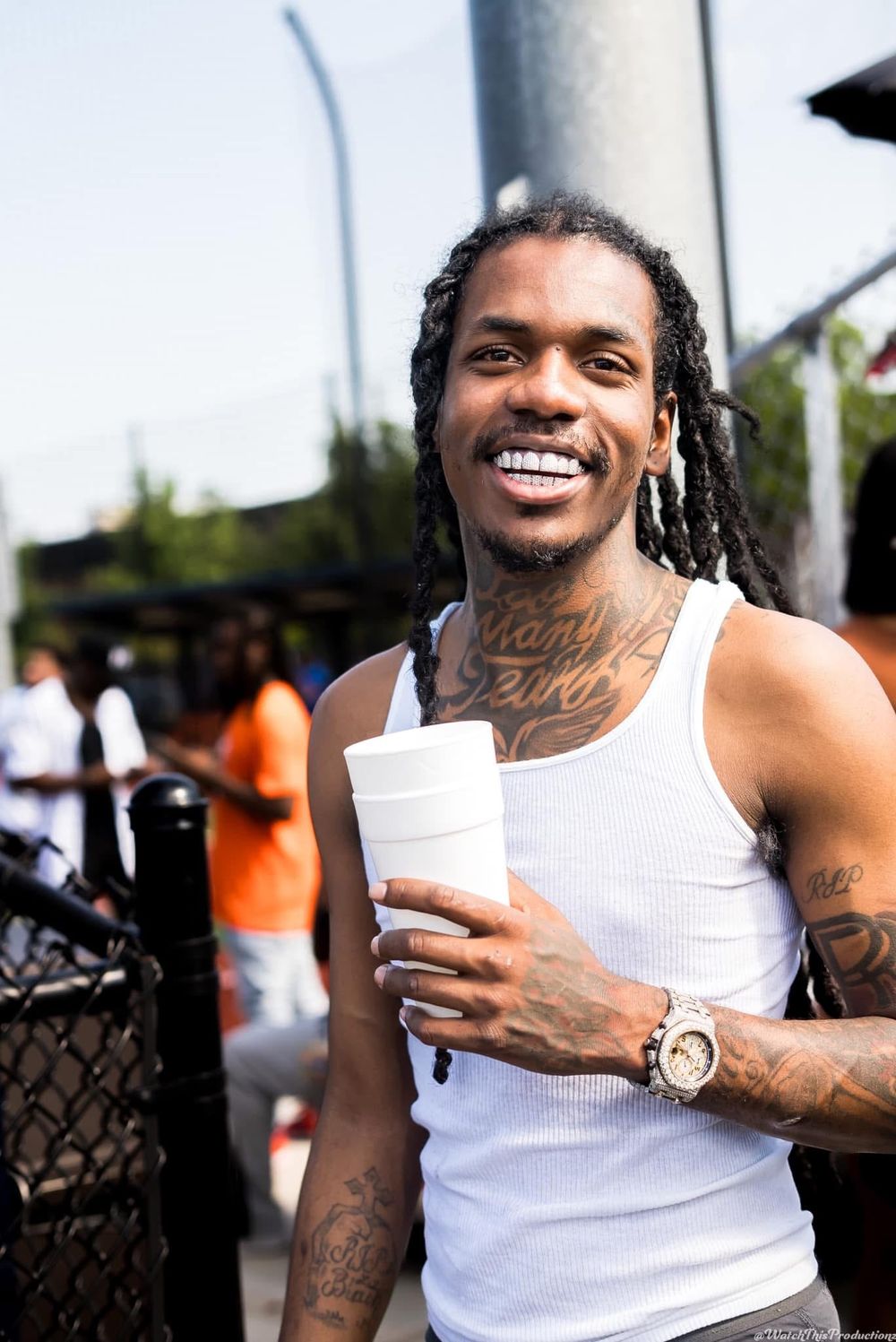 Person in white tank top with tattoos and gold watch smiling outdoors.