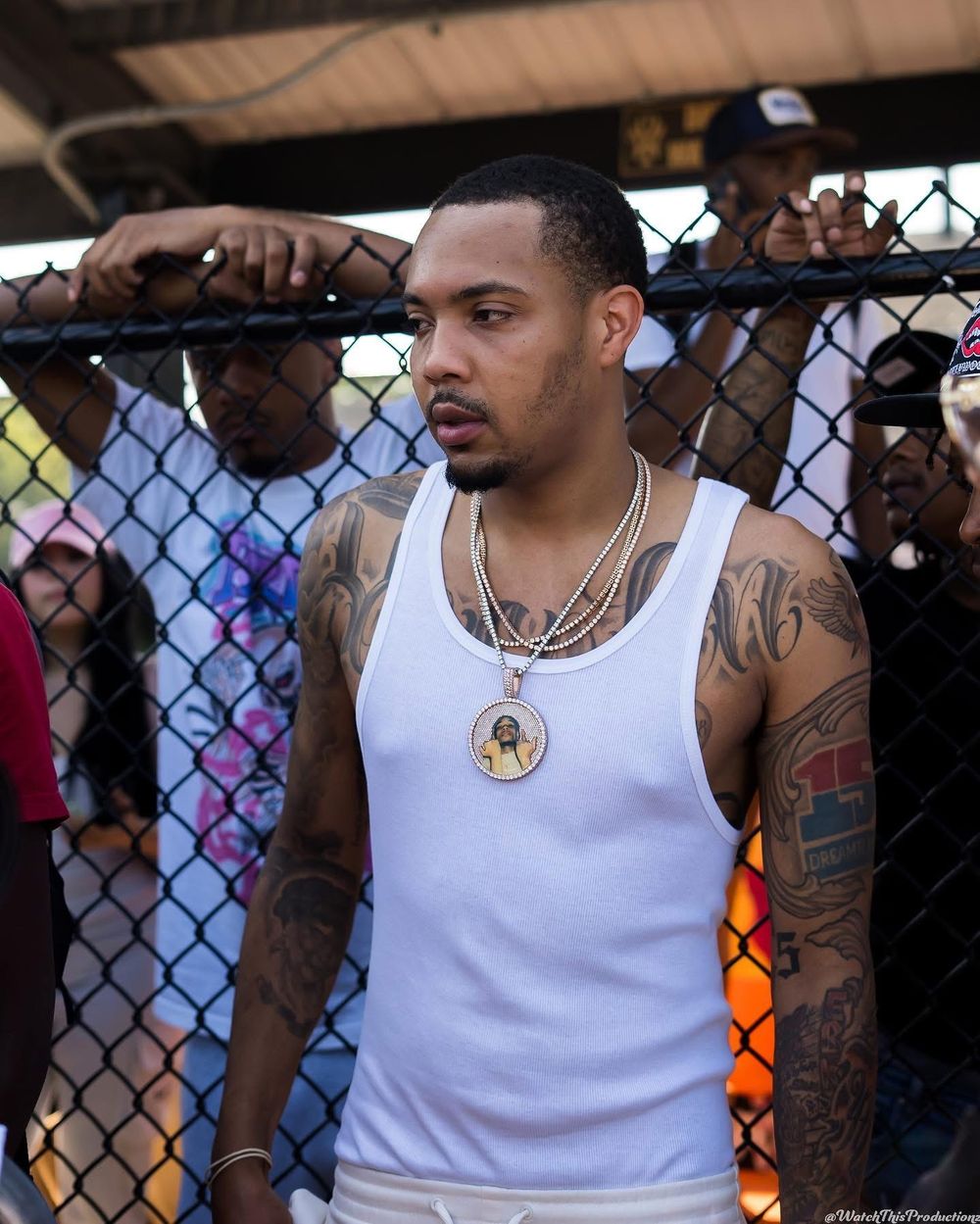 Person wearing white tank top with tattoos and jewelry leaning against chain-link fence.