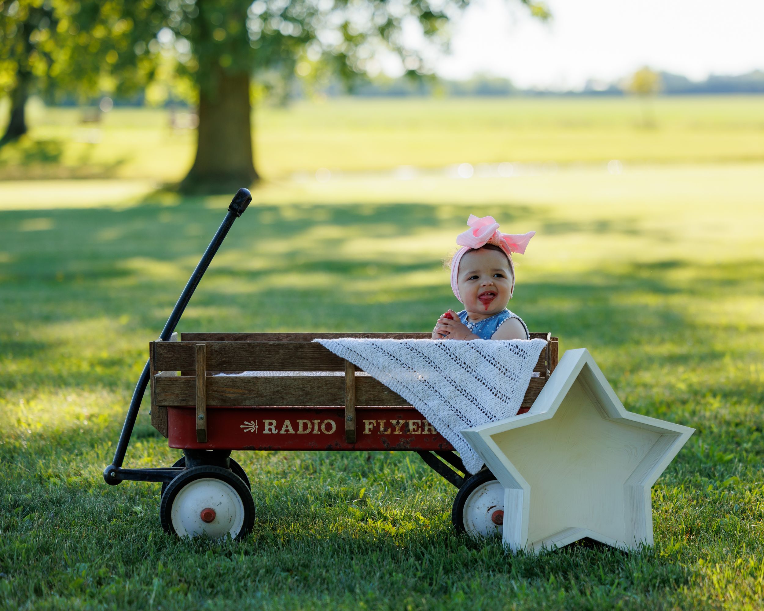 Adorable baby girl in pink bow sitting in a red Radio Flyer wagon beside a star decoration outdoors.