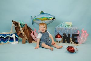Baby boy smiles at first birthday photo shoot with fishing theme, fish balloon, nets, and ONE sign on blue background.