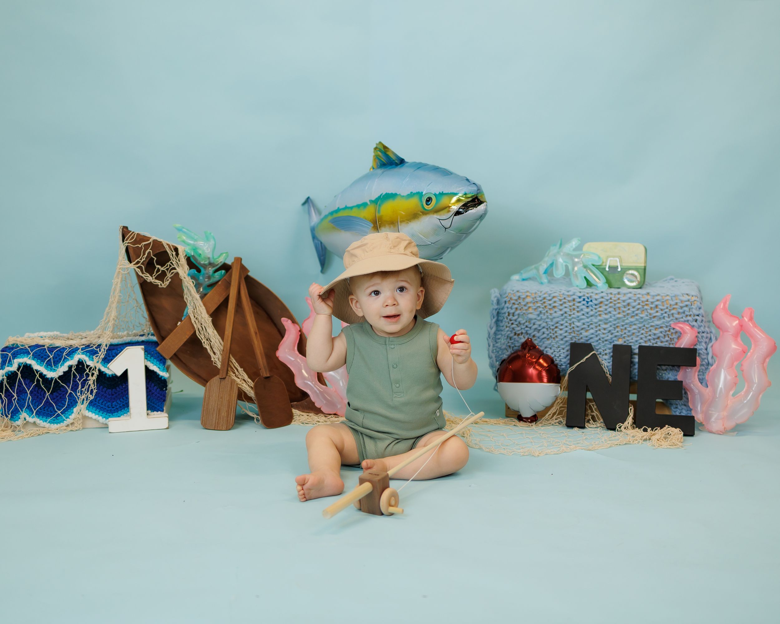Baby's first birthday fishing-themed photo shoot with decorative fish balloon, nets, and ONE sign on blue background.