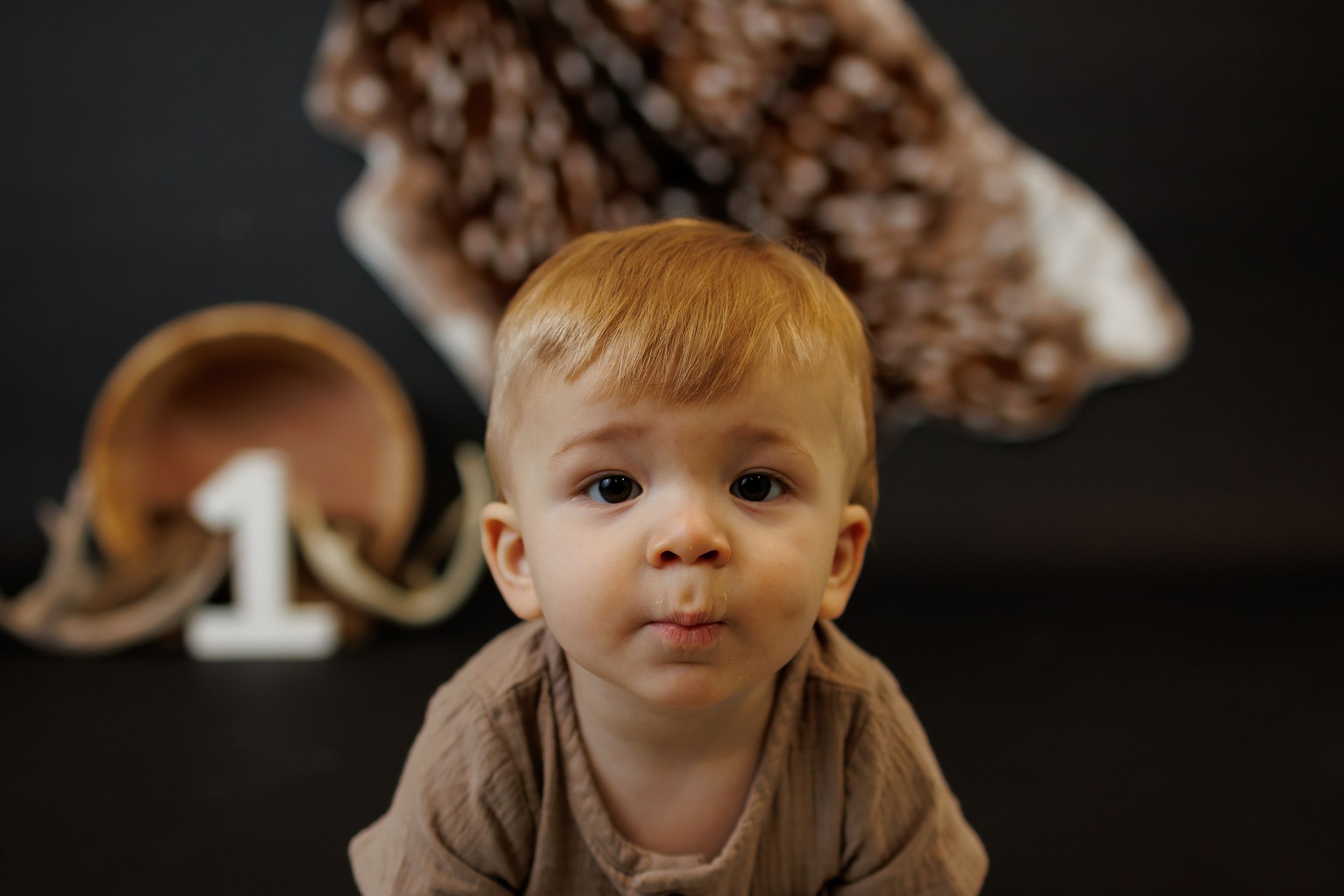 Cute red-haired baby in brown outfit poses near a number one sign and geode for a first birthday photo.