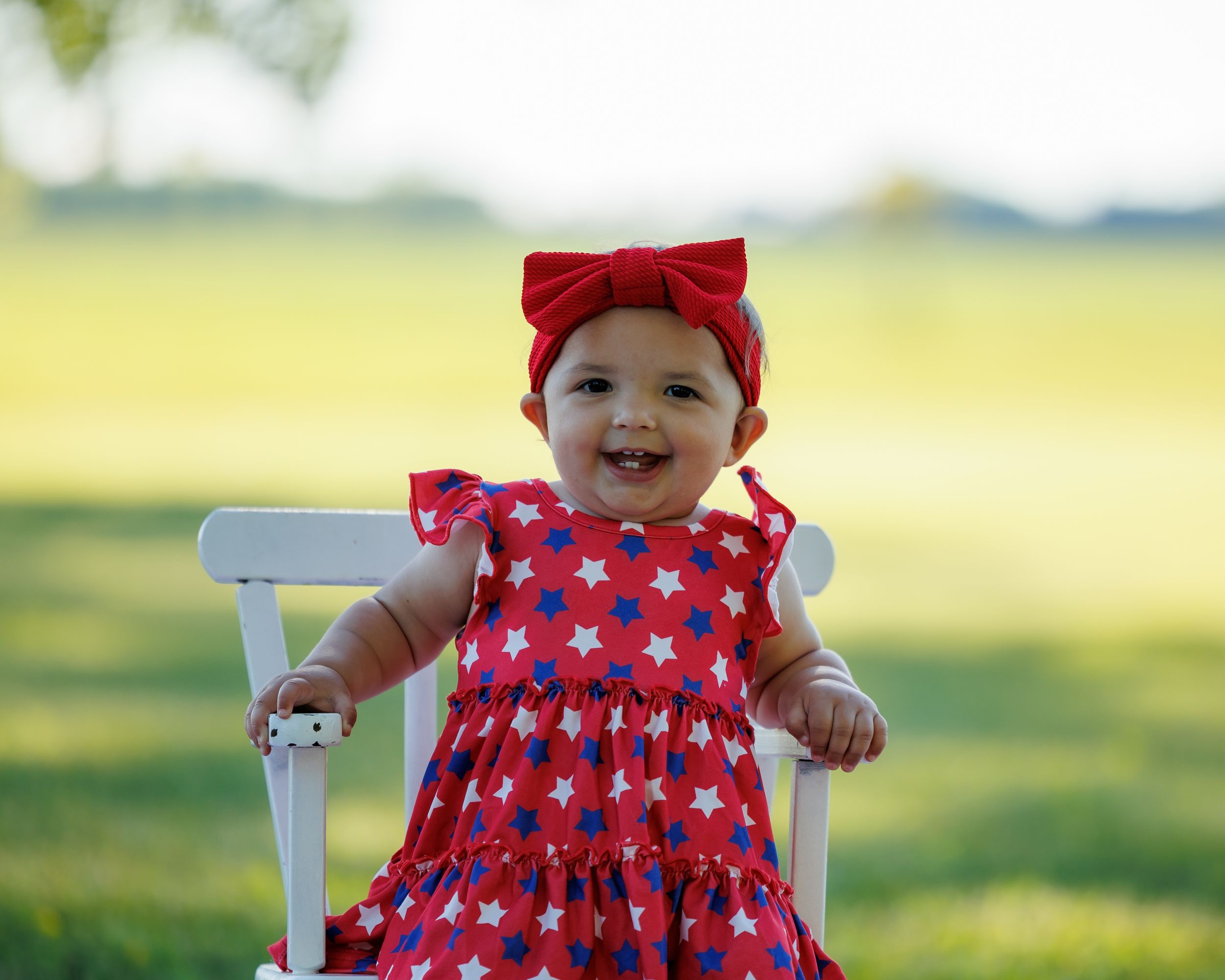 Smiling baby girl in red star-print dress and bow headband sitting in white chair outdoors.