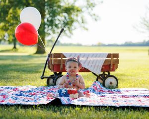 Baby in patriotic outfit sits on American flag quilt with red wagon and red, white balloons outdoors.