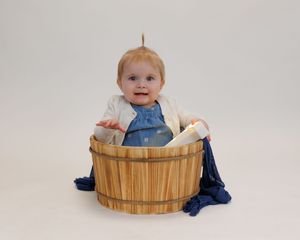 A small child wearing denim sits and plays in a wooden basket against a white background.