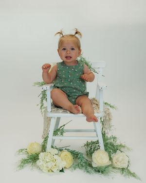 Small baby in green outfit sits in white chair surrounded by yellow roses and greenery creating a whimsical studio portrait.