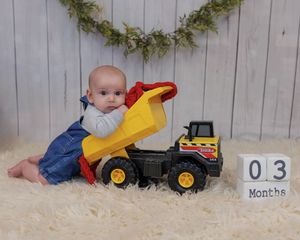 A baby in blue overalls sits on a cream-colored rug with a toy dump truck, with greenery garland on the wall behind them.