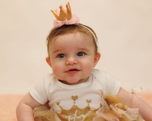 A baby wearing a white onesie with crown design and small crown headpiece smiles while sitting on peach-colored bedding.