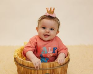 Small baby wearing coral sweatshirt and gold crown sits in wooden bucket during portrait session with cream backdrop.