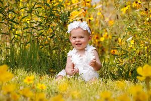 Smiling baby girl in white feather outfit sitting among bright yellow wildflowers in a sunny garden.