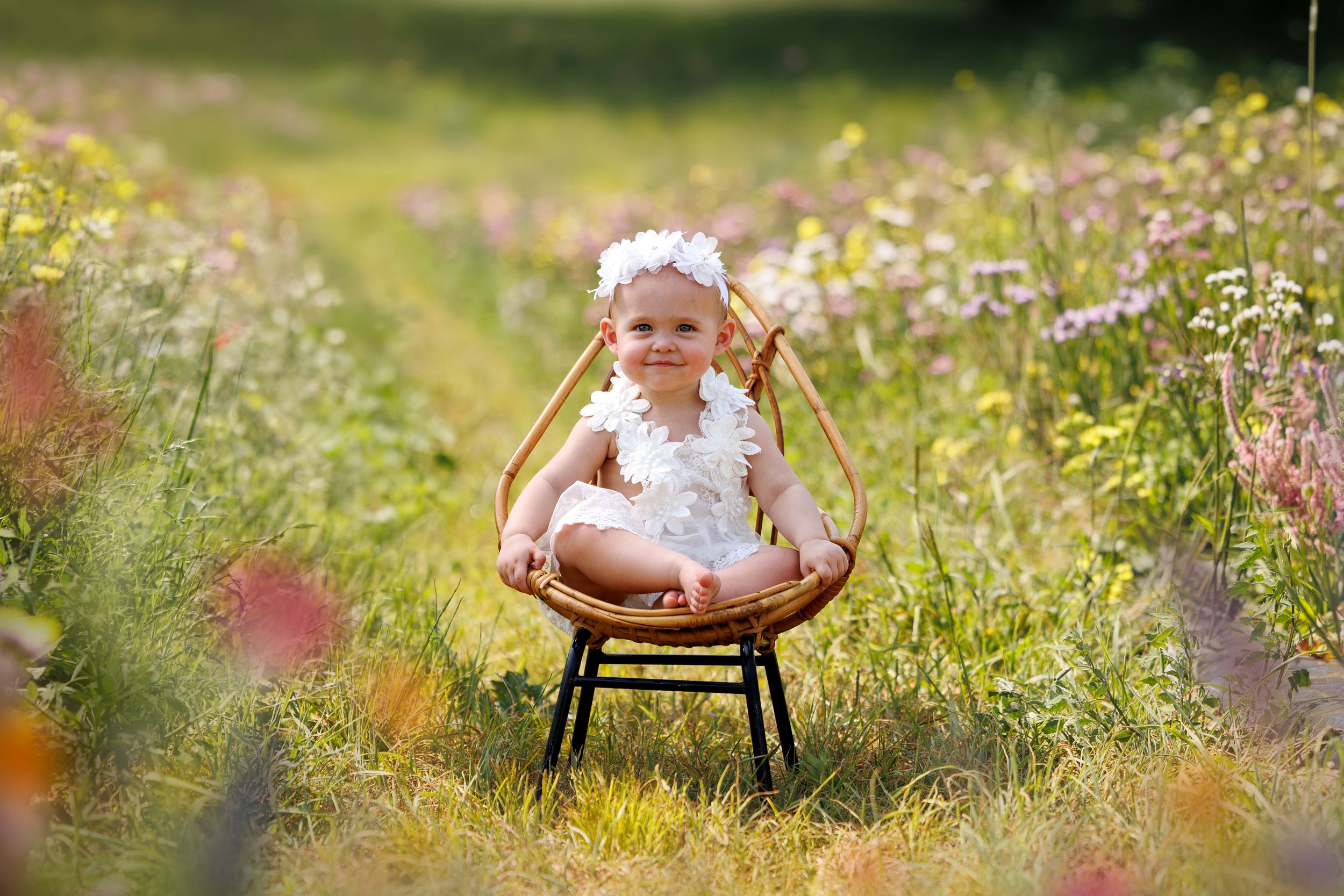 Smiling baby girl in white outfit sitting on wicker chair surrounded by colorful wildflowers in sunny meadow.