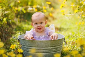 Smiling baby girl in purple dress sitting in a galvanized metal tub surrounded by yellow flowers outdoors.