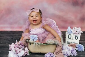 A baby in a pink tutu dress lies in a rustic wooden bowl surrounded by delicate lavender and white flowers.