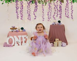 A baby in a purple tulle dress sits on a cream backdrop with floral decorations and stuffed animals.