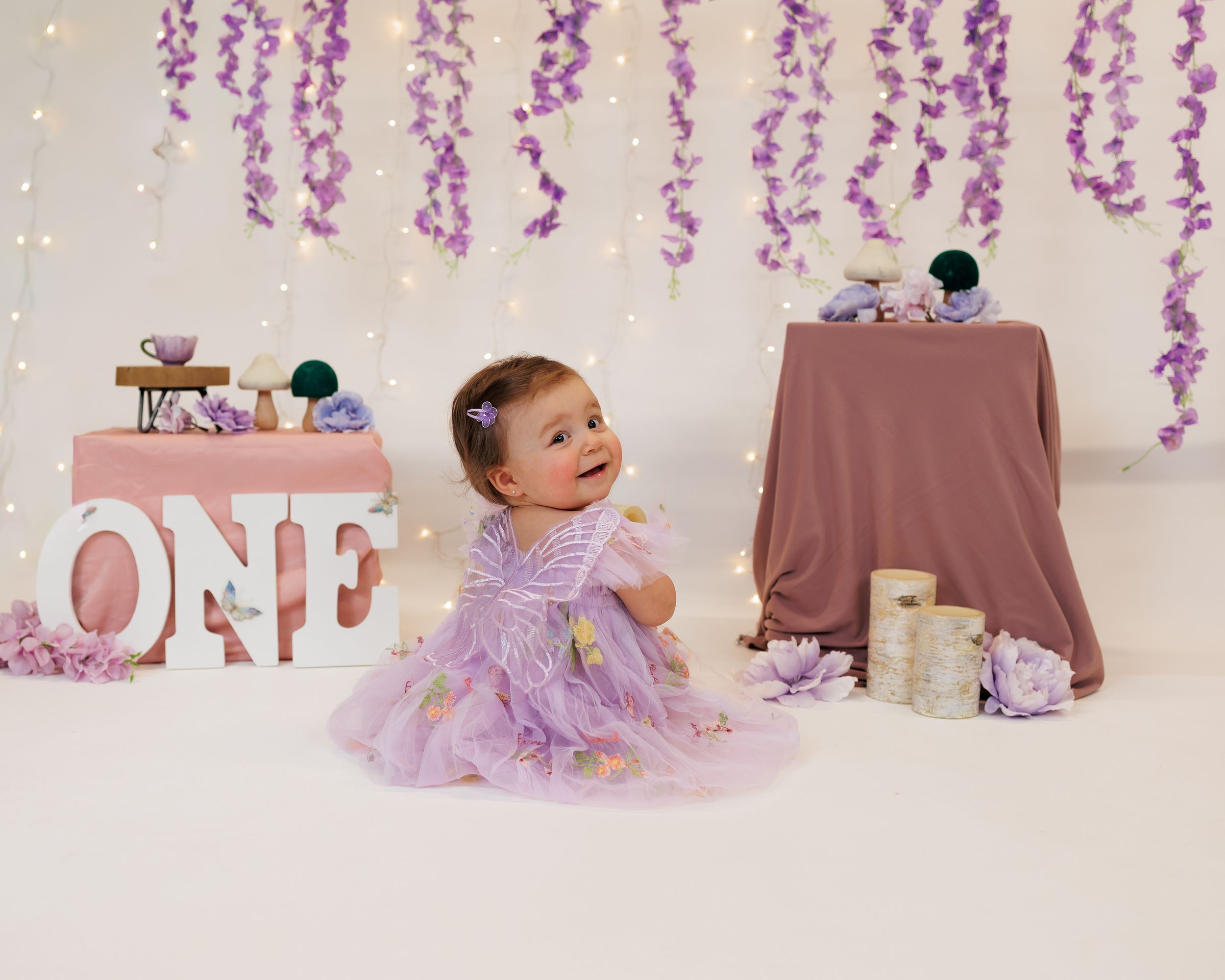 Baby in purple tulle dress sits on floor with 'ONE' letters and hanging purple wisteria decorations for first birthday photoshoot.