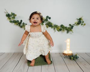 A small child in a white lace dress sits on a green cushion with eucalyptus garland and a glowing candle nearby.