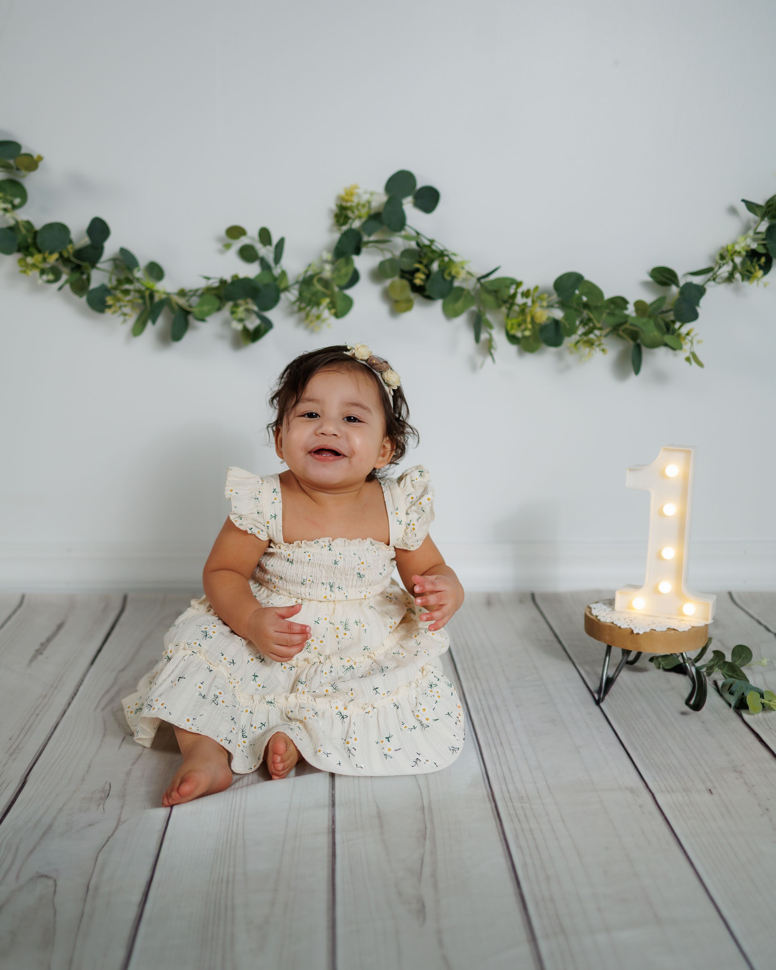 Small child in white dress sitting on wooden floor with greenery garland decoration in background.