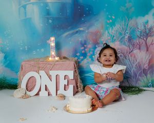 A baby wearing a floral dress sits next to a white birthday cake in a dreamy blue and pink studio setting.