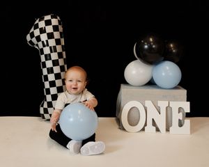 Baby in white outfit sits against checkered backdrop with blue balloons and ONE letters for first birthday photos.