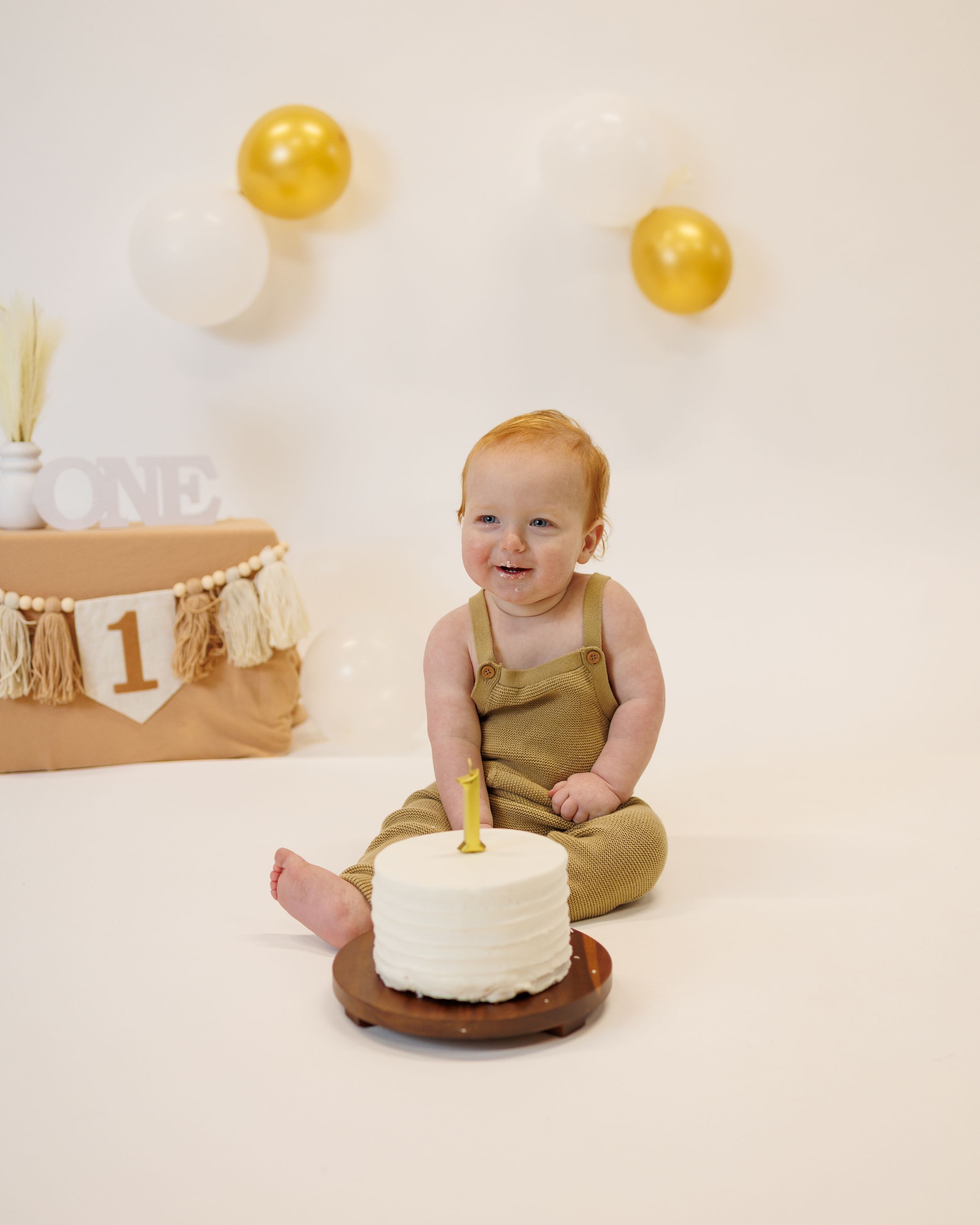 A baby in olive green romper sits on floor with white cake and gold decorations for first birthday celebration.