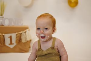 Baby in gold outfit smiles during first birthday photoshoot with white and gold decorations.
