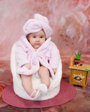 Sweet baby wearing pink plush bathrobe and matching cap sits on circular pink rug near small wooden cabinet with decor.