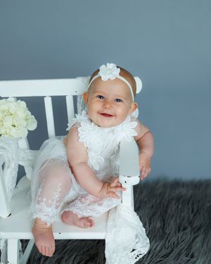 A baby in a delicate white lace dress sits on a plush gray furry rug with white floral decorations nearby.