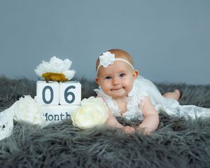 Baby in white lace dress lying on gray rug next to blocks showing 6 months milestone and white flowers.